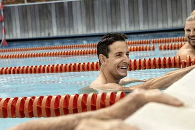 Zwei Männer halten sich am Beckenrand eines Schwimmbeckens fest und lachen. Sie nehmen an einem AOK Gesundheitskurs teil.