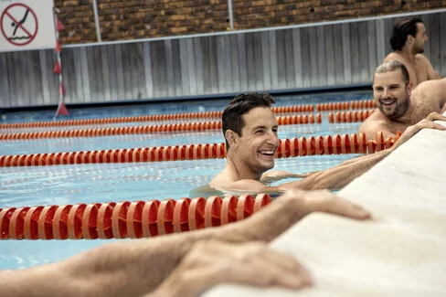 Zwei Männer halten sich am Beckenrand eines Schwimmbeckens fest und lachen. Sie nehmen an einem AOK Gesundheitskurs teil.