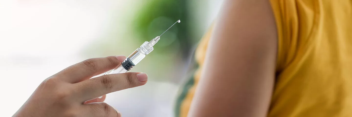 Eine Person hält eine Spritze für eine HPV-Impfung in der Hand, im Hintergrund ist ein junges Mädchen.