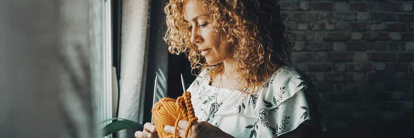 Jüngere Frau mit langen Locken steht an einem Fenster, sie hält eine Sticknadel und gelbe Wolle in der Hand.