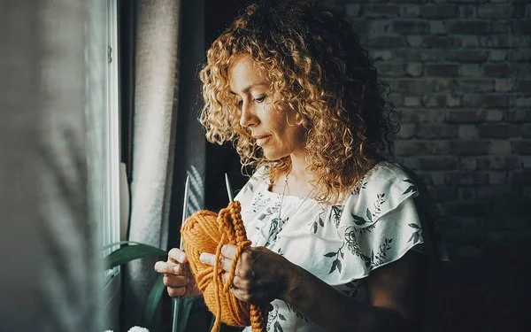 Jüngere Frau mit langen Locken steht an einem Fenster, sie hält eine Sticknadel und gelbe Wolle in der Hand.