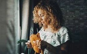 Jüngere Frau mit langen Locken steht an einem Fenster, sie hält eine Sticknadel und gelbe Wolle in der Hand.