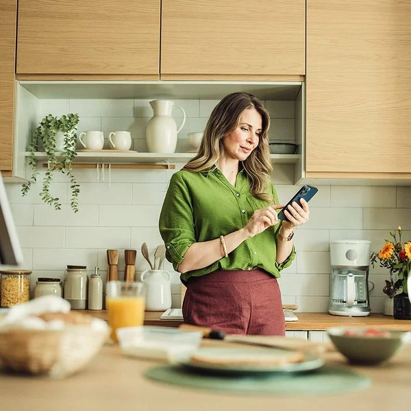Eine Frau mit langen Haaren und in grüner Bluse steht in ihrer Küche und hält ihr Smartphone in der Hand. Sie liest auf dem Display und berührt mit dem Zeigefinger der rechten Hand den Touchscreen.