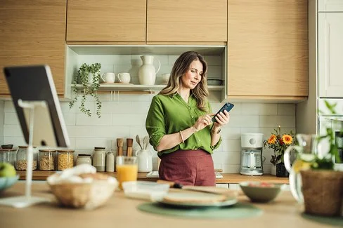 Eine Frau mit langen Haaren und in grüner Bluse steht in ihrer Küche und hält ihr Smartphone in der Hand. Sie liest auf dem Display und berührt mit dem Zeigefinger der rechten Hand den Touchscreen.
