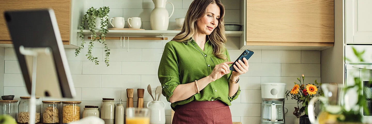 Eine Frau mit langen Haaren und in grüner Bluse steht in ihrer Küche und hält ihr Smartphone in der Hand. Sie liest auf dem Display und berührt mit dem Zeigefinger der rechten Hand den Touchscreen.