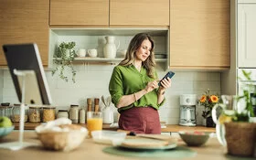 Eine Frau mit langen Haaren und in grüner Bluse steht in ihrer Küche und hält ihr Smartphone in der Hand. Sie liest auf dem Display und berührt mit dem Zeigefinger der rechten Hand den Touchscreen.