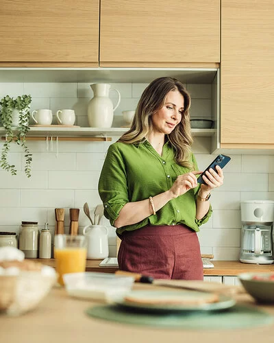 Eine Frau mit langen Haaren und in grüner Bluse steht in ihrer Küche und hält ihr Smartphone in der Hand. Sie liest auf dem Display und berührt mit dem Zeigefinger der rechten Hand den Touchscreen.