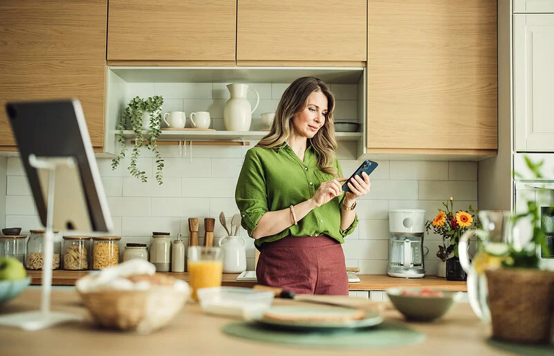 Eine Frau mit langen Haaren und in grüner Bluse steht in ihrer Küche und hält ihr Smartphone in der Hand. Sie liest auf dem Display und berührt mit dem Zeigefinger der rechten Hand den Touchscreen.