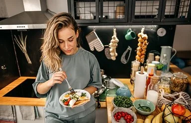 In einer Küche steht eine Frau mit einem Teller in der Hand. Darauf befindet sich ein Salat aus Rucola, Tomaten, Gurken und Eiern sowie Toast. Auf der Arbeitsplatte daneben befinden sich diverse Obst- und Gemüsesorten, Vorratsgläser und Küchenutensilien.