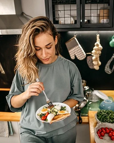 In einer Küche steht eine Frau mit einem Teller in der Hand. Darauf befindet sich ein Salat aus Rucola, Tomaten, Gurken und Eiern sowie Toast. Auf der Arbeitsplatte daneben befinden sich diverse Obst- und Gemüsesorten, Vorratsgläser und Küchenutensilien.