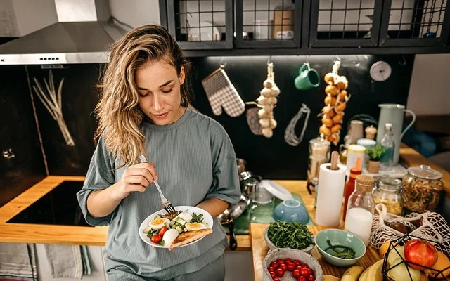 In einer Küche steht eine Frau mit einem Teller in der Hand. Darauf befindet sich ein Salat aus Rucola, Tomaten, Gurken und Eiern sowie Toast. Auf der Arbeitsplatte daneben befinden sich diverse Obst- und Gemüsesorten, Vorratsgläser und Küchenutensilien.