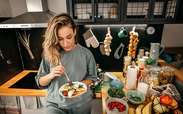 In einer Küche steht eine Frau mit einem Teller in der Hand. Darauf befindet sich ein Salat aus Rucola, Tomaten, Gurken und Eiern sowie Toast. Auf der Arbeitsplatte daneben befinden sich diverse Obst- und Gemüsesorten, Vorratsgläser und Küchenutensilien.