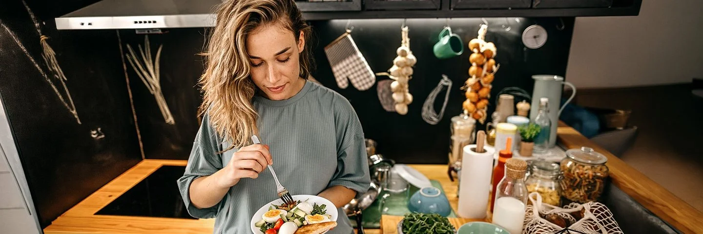 In einer Küche steht eine Frau mit einem Teller in der Hand. Darauf befindet sich ein Salat aus Rucola, Tomaten, Gurken und Eiern sowie Toast. Auf der Arbeitsplatte daneben befinden sich diverse Obst- und Gemüsesorten, Vorratsgläser und Küchenutensilien.