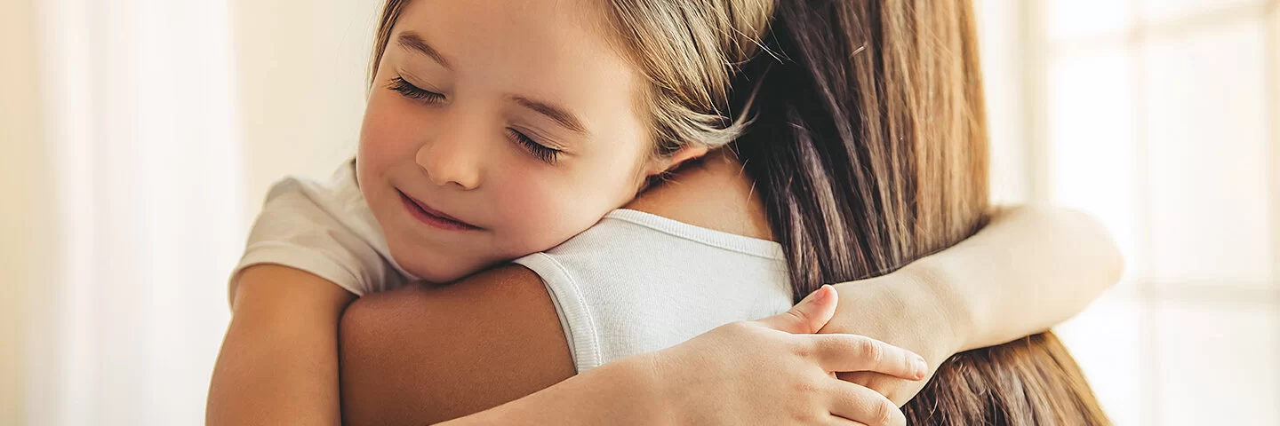 Mutter trägt Tochter mit Wachstumsschmerzen auf dem Arm, die Tochter umarmt die Mutter mit geschlossenen Augen und lächelt.