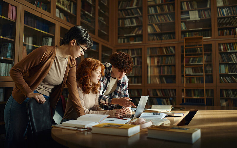 In einer alten Bibliothek ist eine Gruppe Studierender an einem Tisch mit Laptop, Büchern und Notizen in ein Gespräch vertieft.