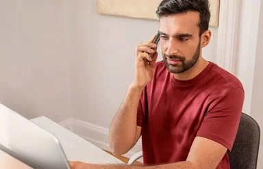 Mann mit rotem T-Shirt sitzt an einem Laptop und telefoniert in einem hellen Raum