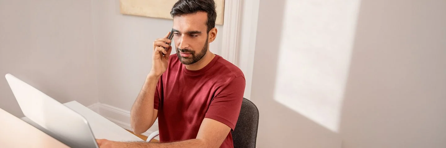Mann mit rotem T-Shirt sitzt an einem Laptop und telefoniert in einem hellen Raum