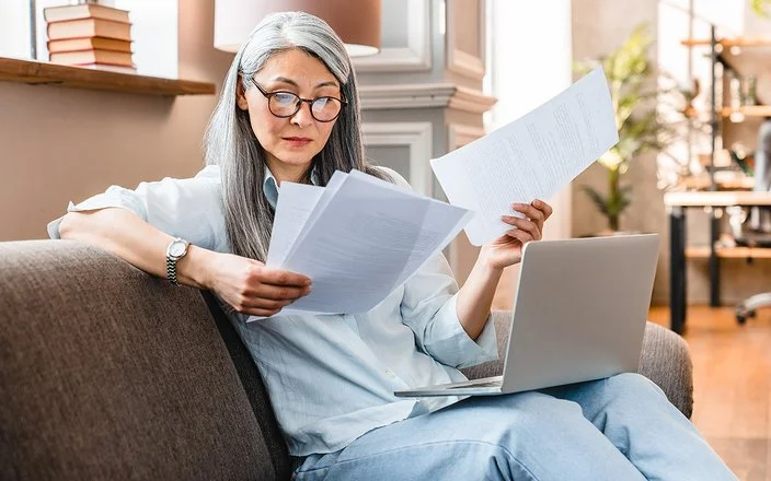 Eine Frau mittleren Alters sitzt mit einem Laptop auf dem Schoß auf einem Sofa und hält mehrere Zettel in den Händen.
