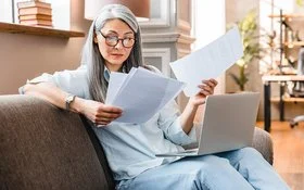 Eine Frau mittleren Alters sitzt mit einem Laptop auf dem Schoß auf einem Sofa und hält mehrere Zettel in den Händen.