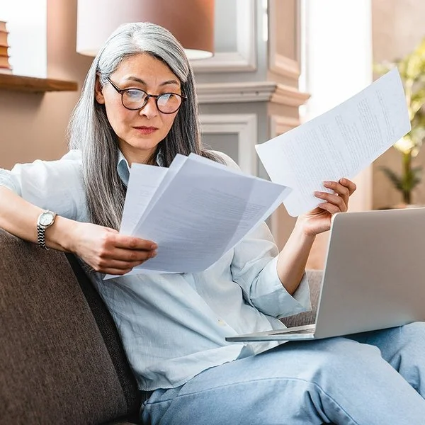 Eine Frau mittleren Alters sitzt mit einem Laptop auf dem Schoß auf einem Sofa und hält mehrere Zettel in den Händen.