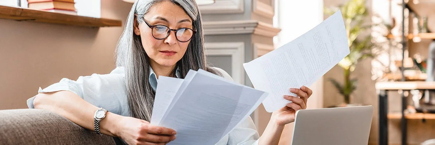 Eine Frau mittleren Alters sitzt mit einem Laptop auf dem Schoß auf einem Sofa und hält mehrere Zettel in den Händen.