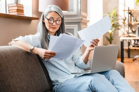Eine Frau mittleren Alters sitzt mit einem Laptop auf dem Schoß auf einem Sofa und hält mehrere Zettel in den Händen.