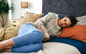 Eine Frau im Bett liegt mit angewinkelten Beinen auf der linken Seite und hält die Arme schützend auf ihren Bauch.