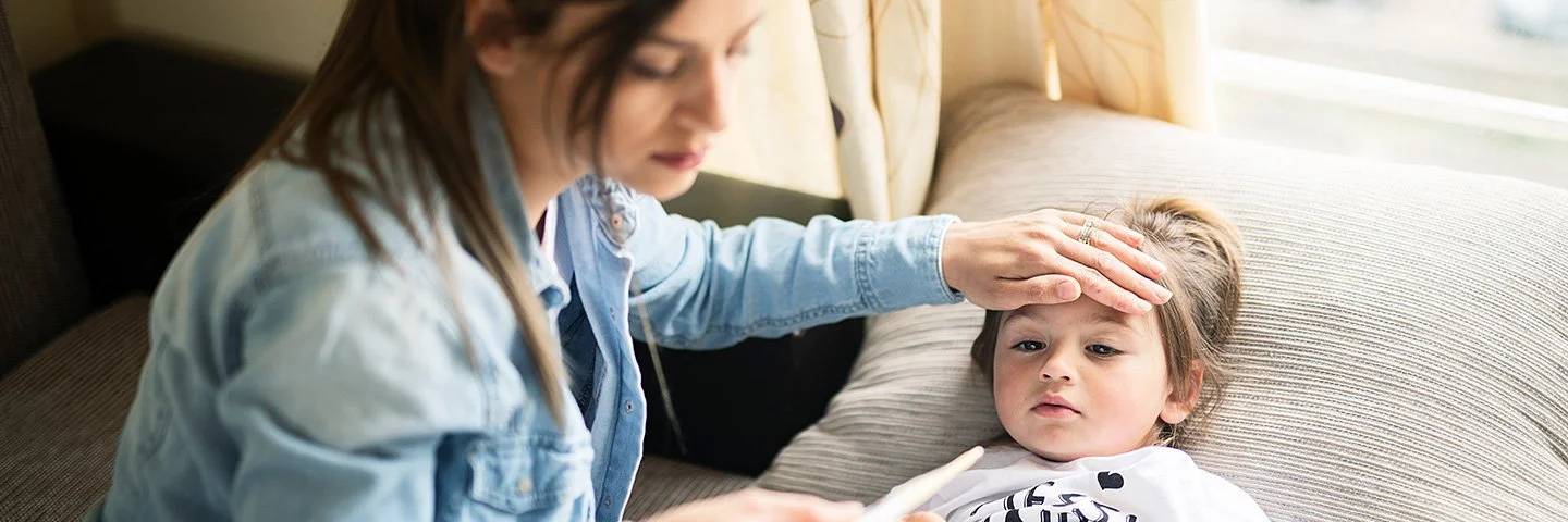 Eine Mutter misst die Temperatur ihrer kleinen Tochter, die auf einem Sofa liegt und mit einer Decke zugedeckt ist. Sie legt dem Kind die linke Hand auf die Stirn. In der rechten Hand hält sie ein Fieberthermometer.