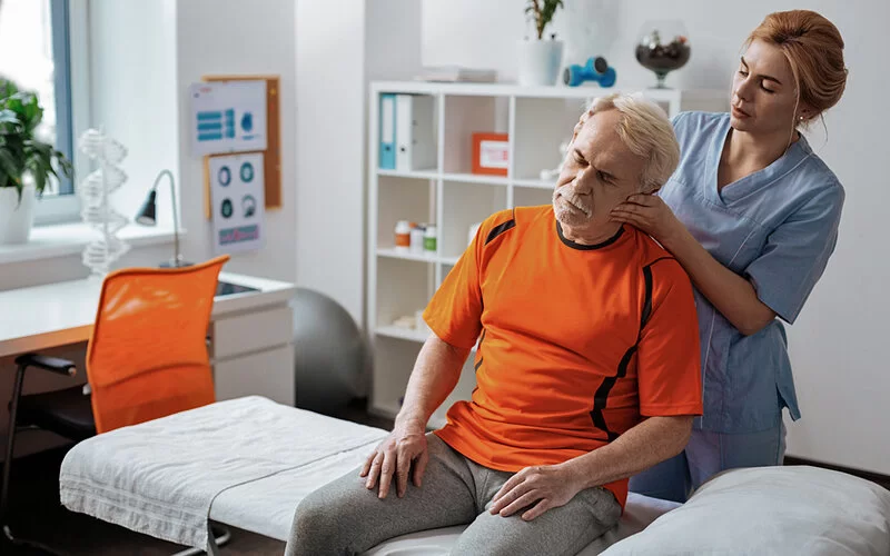 Ein älterer Patient sitzt auf einer Liege, während eine Ärztin seinen Kopf mitsamt Oberkörper leicht nach links bewegt.