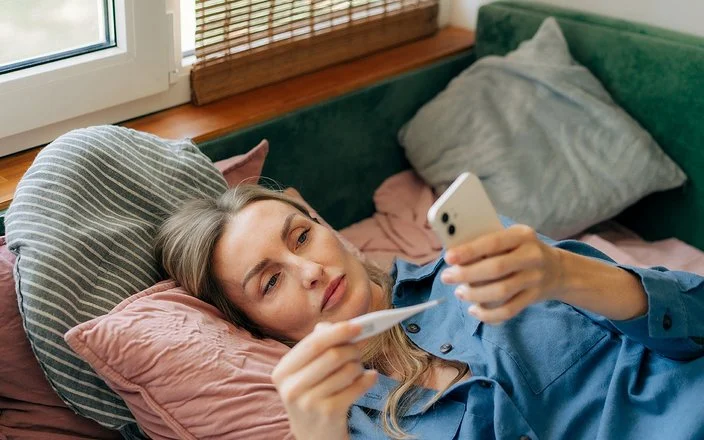 Eine jüngere Frau liegt auf der Couch, hält ein Thermometer in der einen Hand und schaut auf ihr Smartphone, das sie in der anderen Hand hält.