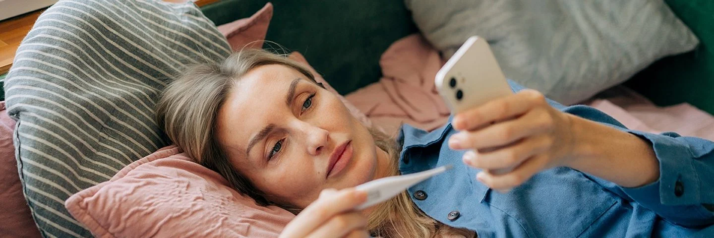 Eine jüngere Frau liegt auf der Couch, hält ein Thermometer in der einen Hand und schaut auf ihr Smartphone, das sie in der anderen Hand hält.