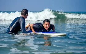 Mädchen liegt auf einem Surfbrett und hat sichtlich Spaß dabei. Ein Surflehrer hält das Brett fest und hilft ihr beim Surfen lernen.