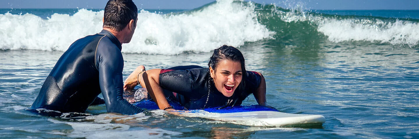 Mädchen liegt auf einem Surfbrett und hat sichtlich Spaß dabei. Ein Surflehrer hält das Brett fest und hilft ihr beim Surfen lernen.