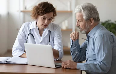Eine Ärztin in weißem Kittel zeigt einem Patienten etwas am Bildschirm ihres Laptops.