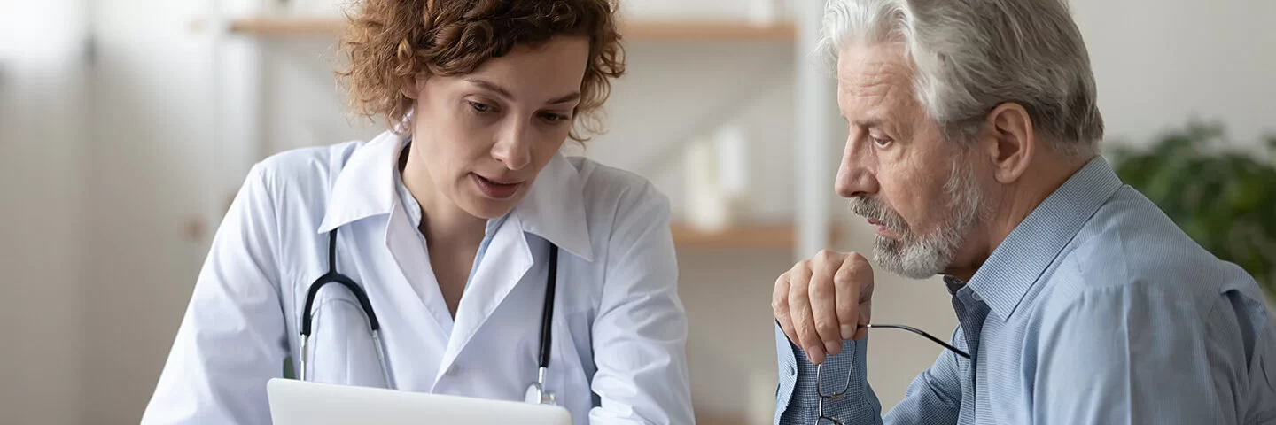 Eine Ärztin in weißem Kittel zeigt einem Patienten etwas am Bildschirm ihres Laptops.