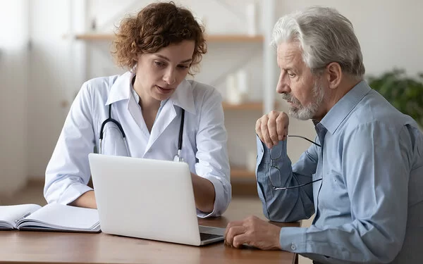 Eine Ärztin in weißem Kittel zeigt einem Patienten etwas am Bildschirm ihres Laptops.