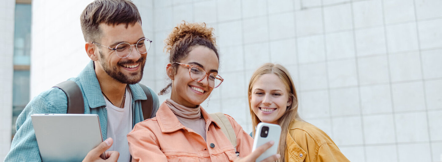 Ein männlicher und zwei weibliche Studierende stehen gemeinsam im Freien und schauen lächelnd auf ein Smartphone.