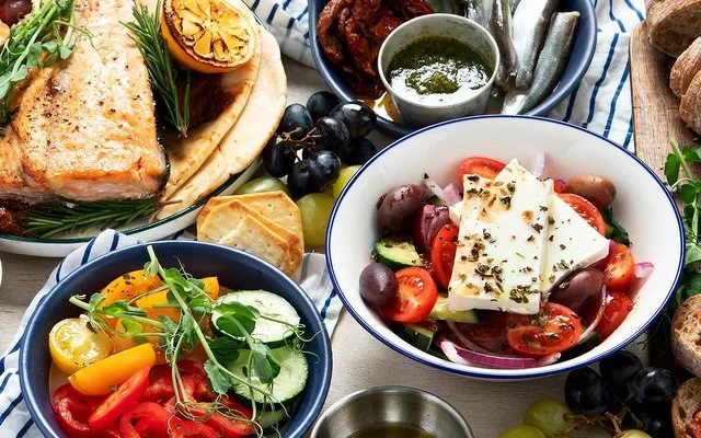 Aufsicht auf einen Tisch, auf dem verschiedene Lebensmittel wie Gemüse, Trauben, Oliven, Hirtensalat, Brot und Fisch in Tellern und Schalen angerichtet sind.