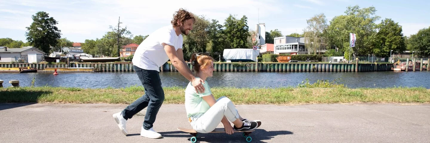 Eine Frau sitzt auf einem Skateboard und wird von einem Mann am Rücken angeschoben.