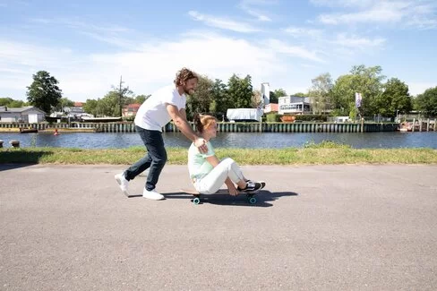 Eine Frau sitzt auf einem Skateboard und wird von einem Mann am Rücken angeschoben.