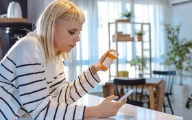Eine junge Frau mit blonden Haaren und einem weiß-schwarz gestreiften Langarmshirt betrachtet eine orange Medikamentendose. Dabei hält sie ein Smartphone in der Hand und stützt sich auf der Küchenarbeitsplatte ab.