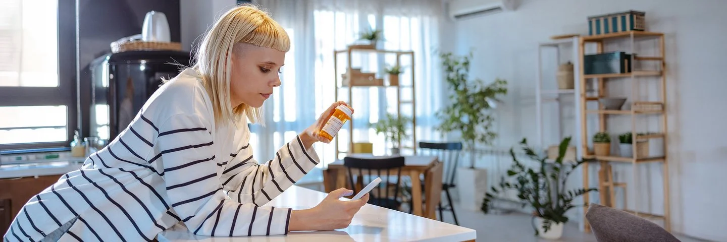 Eine junge Frau mit blonden Haaren und einem weiß-schwarz gestreiften Langarmshirt betrachtet eine orange Medikamentendose. Dabei hält sie ein Smartphone in der Hand und stützt sich auf der Küchenarbeitsplatte ab.