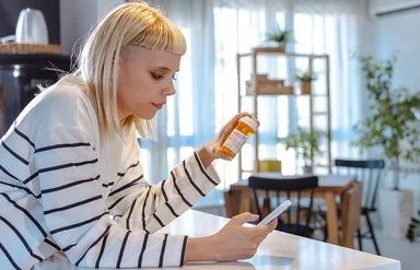 Eine junge Frau mit blonden Haaren und einem weiß-schwarz gestreiften Langarmshirt betrachtet eine orange Medikamentendose. Dabei hält sie ein Smartphone in der Hand und stützt sich auf der Küchenarbeitsplatte ab.