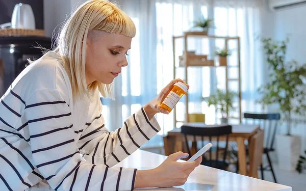 Eine junge Frau mit blonden Haaren und einem weiß-schwarz gestreiften Langarmshirt betrachtet eine orange Medikamentendose. Dabei hält sie ein Smartphone in der Hand und stützt sich auf der Küchenarbeitsplatte ab.