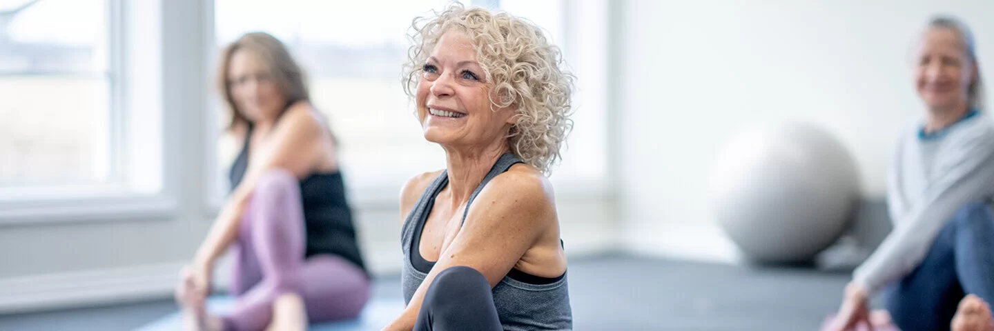 Eine ältere Frau sitzt auf einer Yoga-Matte und lächelt.