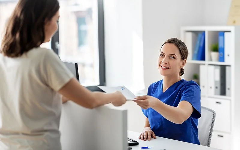 Eine Ärztin sitzt an einem Schreibtisch im Behandlungszimmer und überreicht einer Patientin ein Dokument.