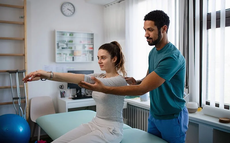 Eine junge Frau sitzt mit nach vorn gestreckten Armen auf einer Behandlungsliege. Hinter ihr steht ein Physiotherapeut, der ihre Haltung korrigiert.