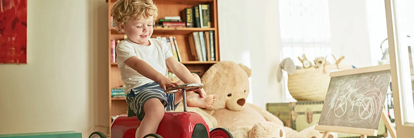 Ein Junge sitzt auf einem Spielzeugauto in seinem Zimmer, das voll mit Spielsachen ist.