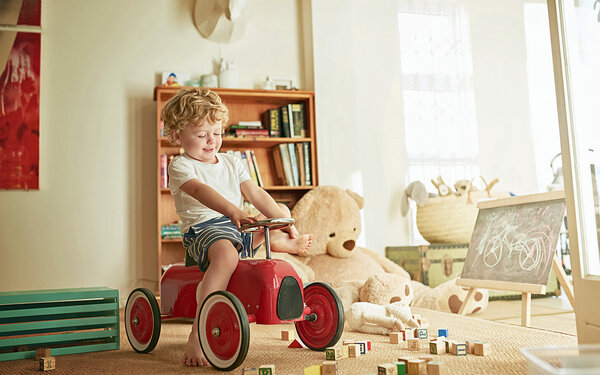 Ein Junge sitzt auf einem Spielzeugauto in seinem Zimmer, das voll mit Spielsachen ist.