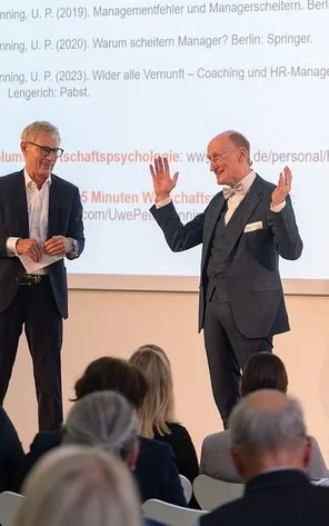 Zwei Männer stehen auf einer Bühne vor einer Gruppe Menschen.